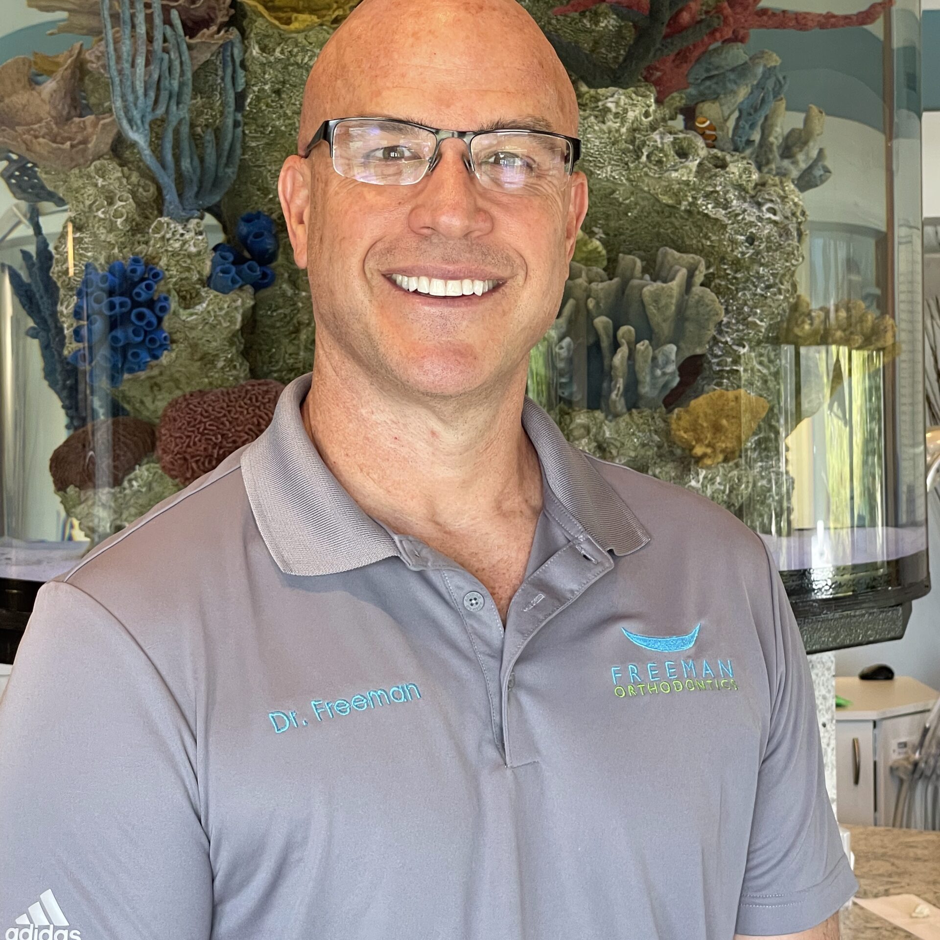 Dr. Freeman smiling in front of an aquarium, wearing a gray polo with the Freeman Orthodontics logo.